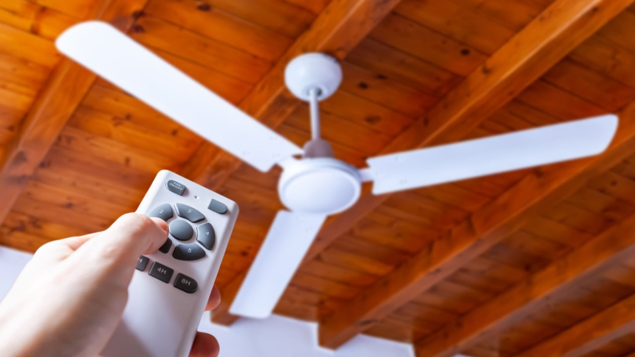 A man uses a remote control to turn on a white ceiling fan mounted in a house with wooden ceilings