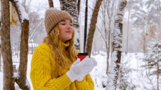 a woman in a yellow sweater holds a coffee cup in the middle of a snowy woods