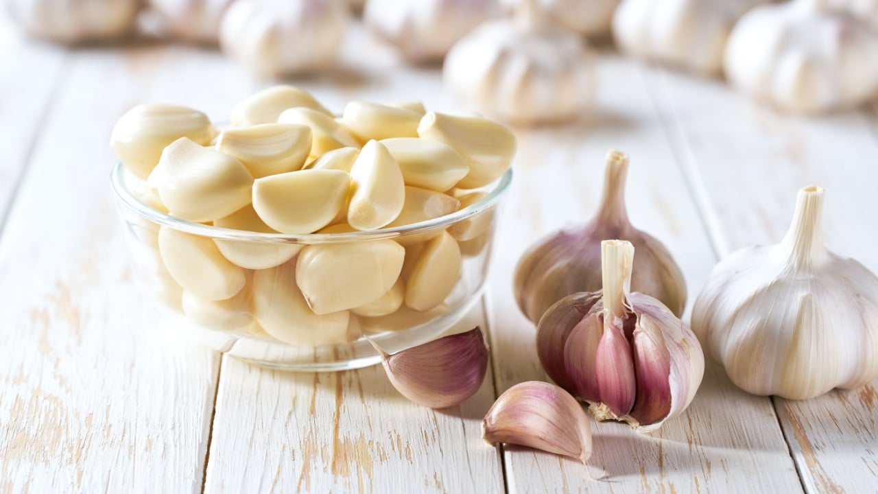 Garlic cloves in a glass bowl with peeled garlic on a white wooden table