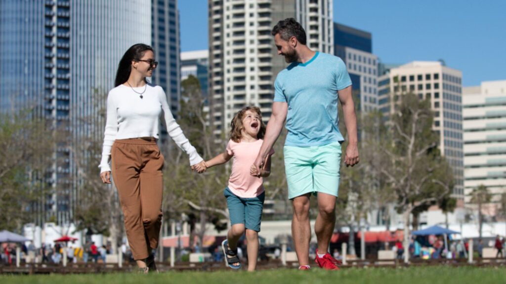 happy young family with couple and child walking in grass park in city
