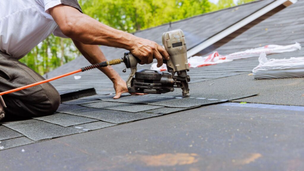 In addition to using an air pneumatic nail gun, roofer installs new asphalt bitumen shingles