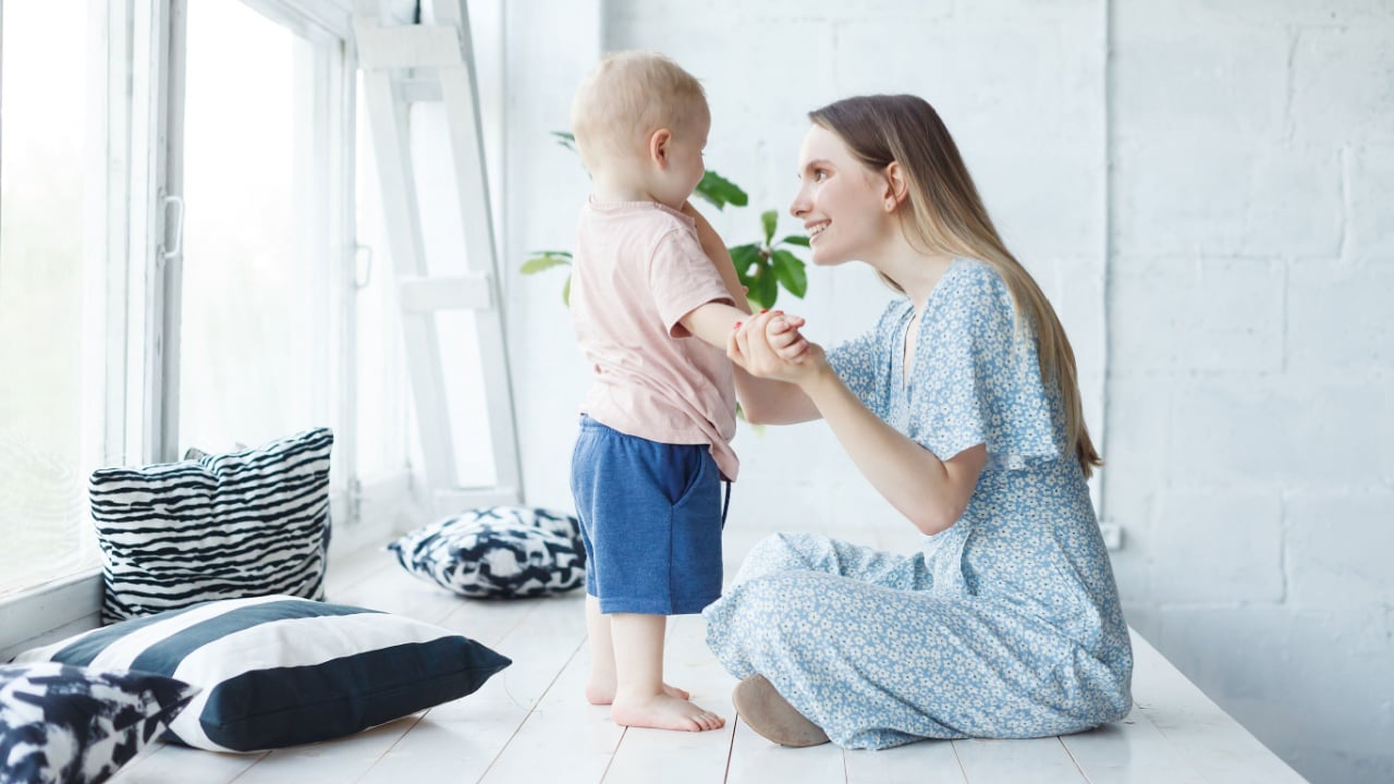 Young, caring mother looks at her baby with great love and warmth, and she is happy in motherhood. Mom adores her small baby, sits over cozy fashionable Scandinavian home interior with big window
