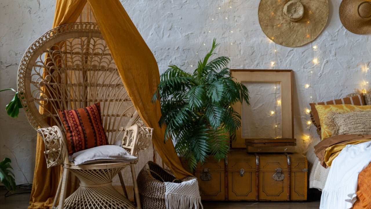Quiet bohemian room with cozy interior, wicker chair, pillows, cushions, green plants in flower pot, bed and brimmed hats on white brick wall