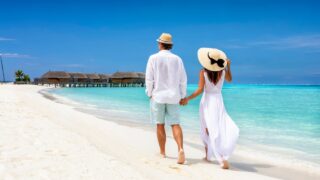 Happy couple in white clothing and with hats walks down a tropical beach with turquoise sea in the Maldives islands