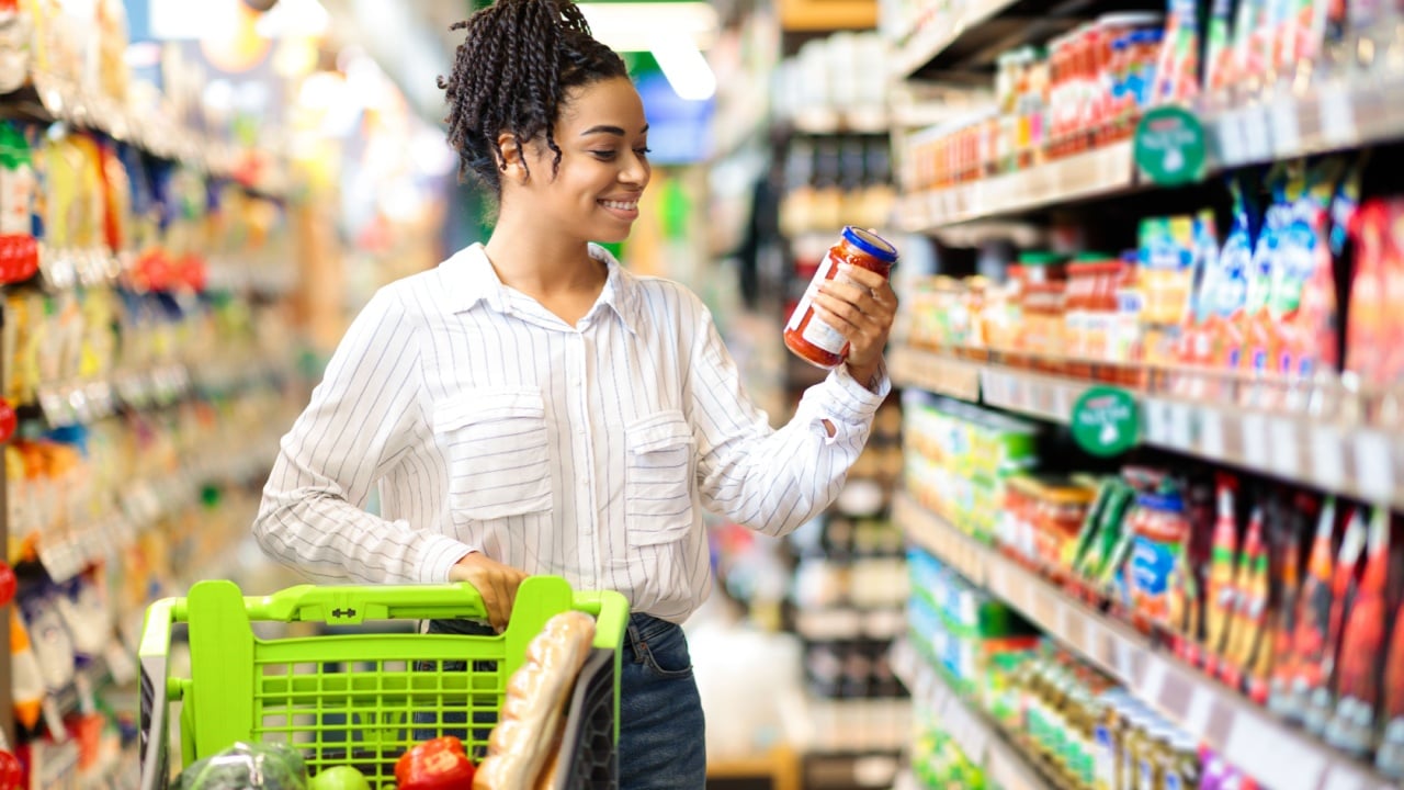 Happy Black Woman Buyer Doing Groceries Shopping Choosing Food And Buying Products Standing With Shop Cart Near Shelf In Modern Supermarket Indoors.