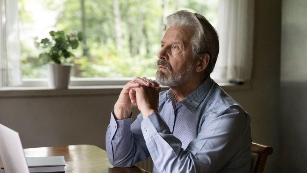 thinking mature grey haired 60s - 70s aged man sitting at table with laptop at home, looking away lost focus. Thoughtful retired OAP working at computer from home, thinking over retirement during break