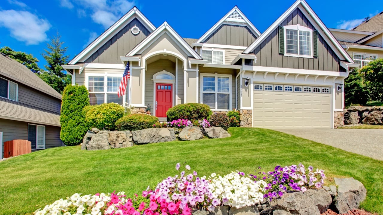 Grey house exterior with entrance porch and red door. Beautiful front yard landscape with vivid flower and stones