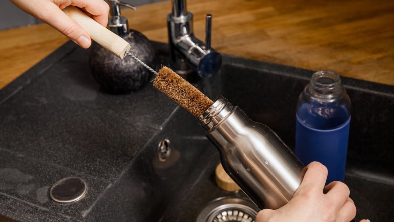 Washing cleaning reusable drinking bottle with coconut fiber bottle cleaning brush in home kitchen. Sustainable lifestyle concept.