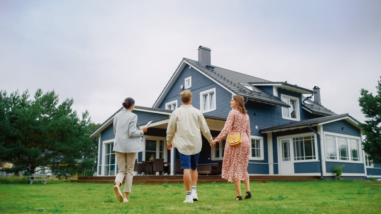 Real Estate Agent Showing a Beautiful Big House to a Young Successful Couple. People Standing Outside on a Warm Day on a Lawn, Talking with Businesswoman, Discussing Buying a New Home.