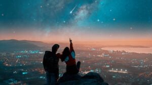 silhouettes of a couple sitting on the top of the mountain looking and pointing out at shooting star and milky way over the city lights on the horizon