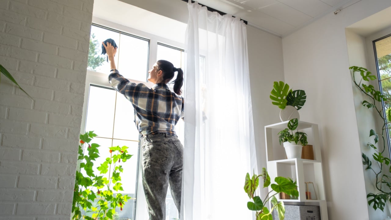 Woman manually washes the window of the house with a rag with spray cleaner and mop inside the interior with white curtains. Restoring order and cleanliness in the spring, cleaning servise