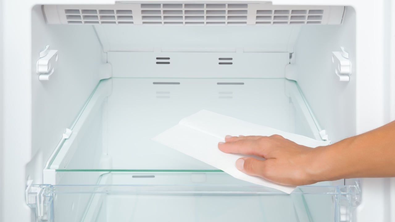 Young adult woman hand wiping glass shelf of refrigerator with dry white paper napkin. Closeup.