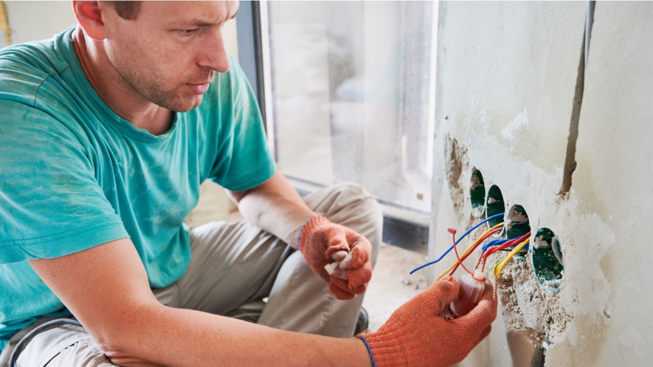 Close up of man in work gloves sitting by the wall and putting electrical wire cable in bottle with liquid. Male electrician installing electrical wiring in apartment under renovation.