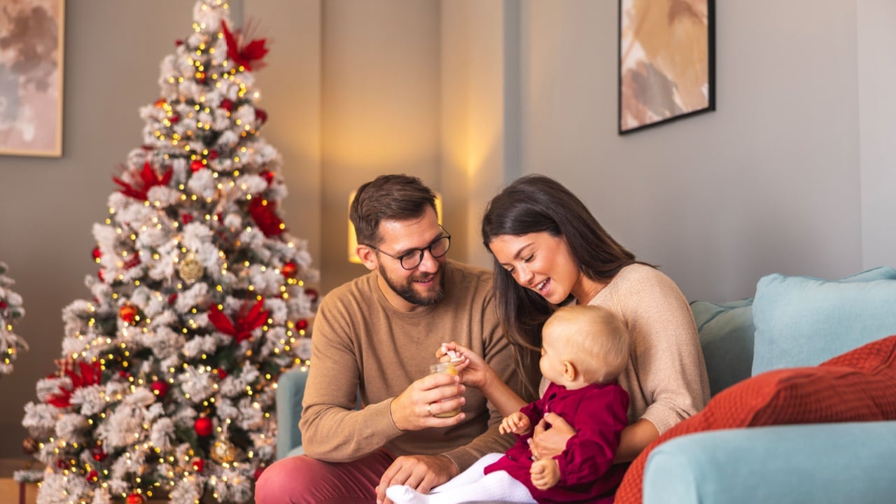 Young parents sitting on the couch by the Christmas tree, feeding baby while spending Christmas day together at home