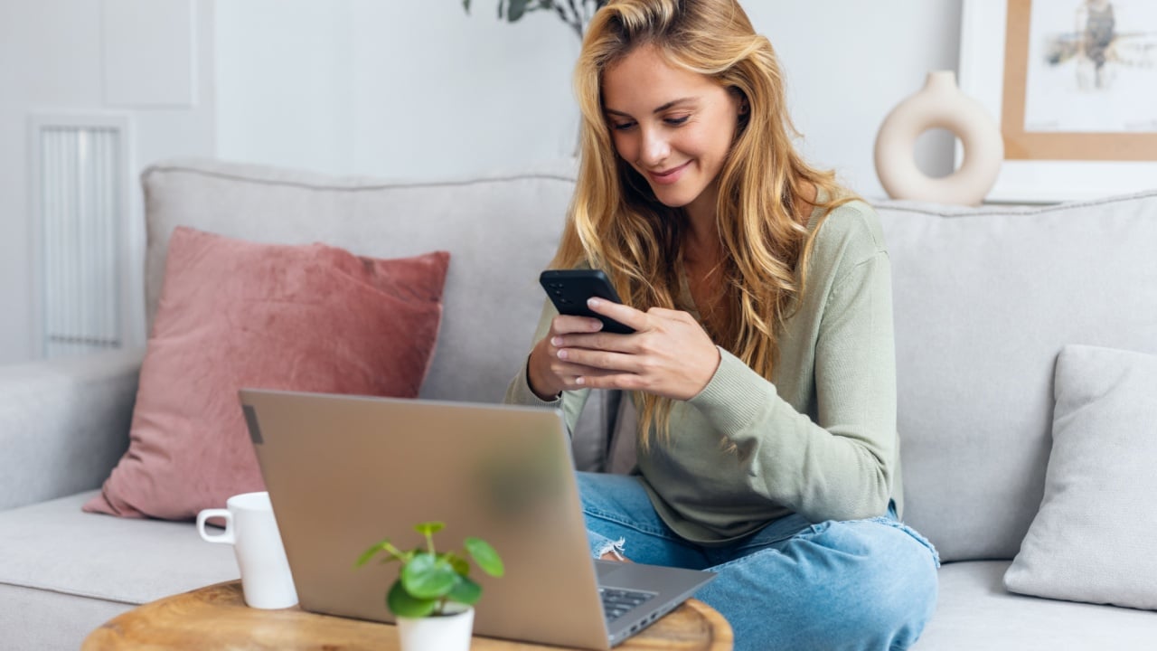 Shot of beautiful woman working with her laptop while using mobile phone sitting on a couch at home.