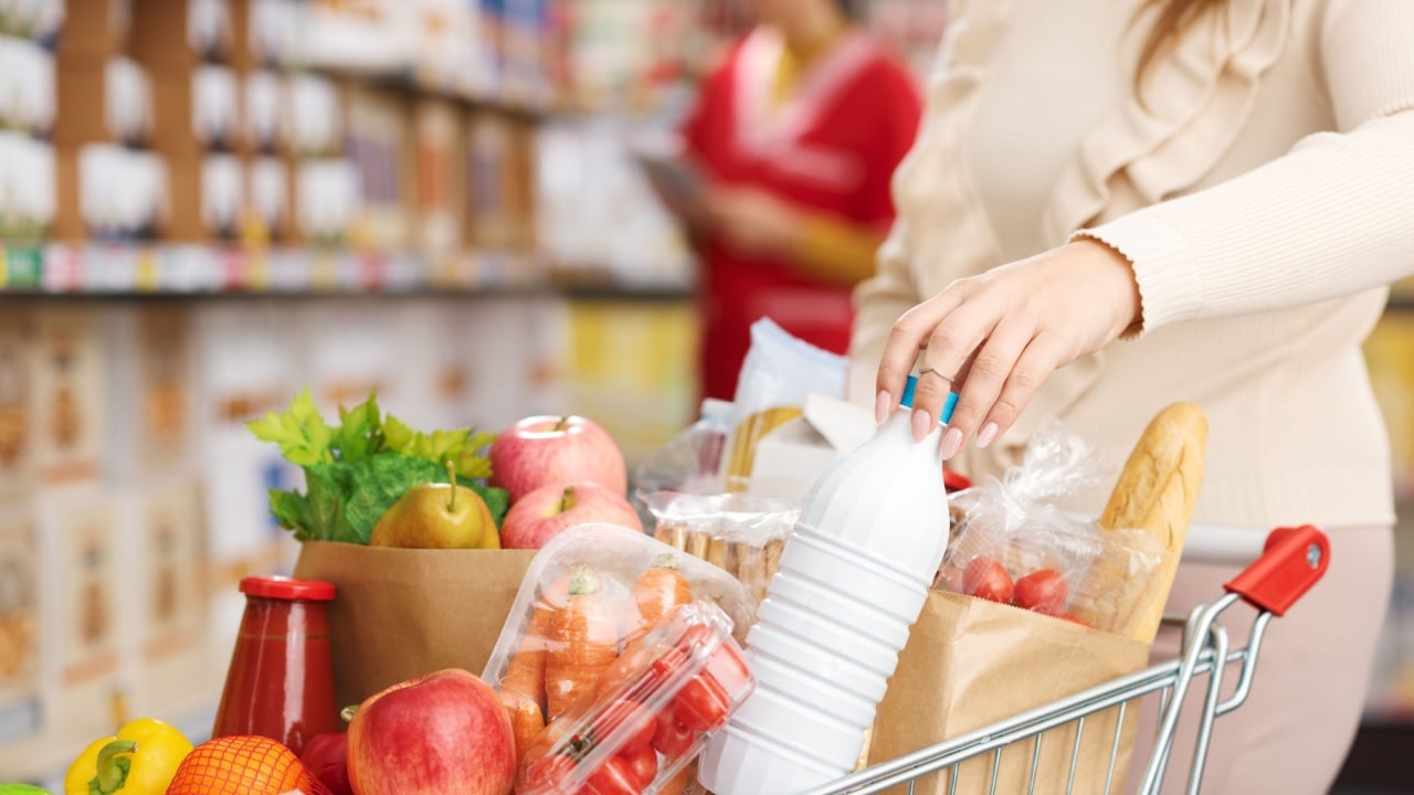 Woman buying fresh groceries at the supermarket, she is pushing a full shopping cart and holding a bottle of milk