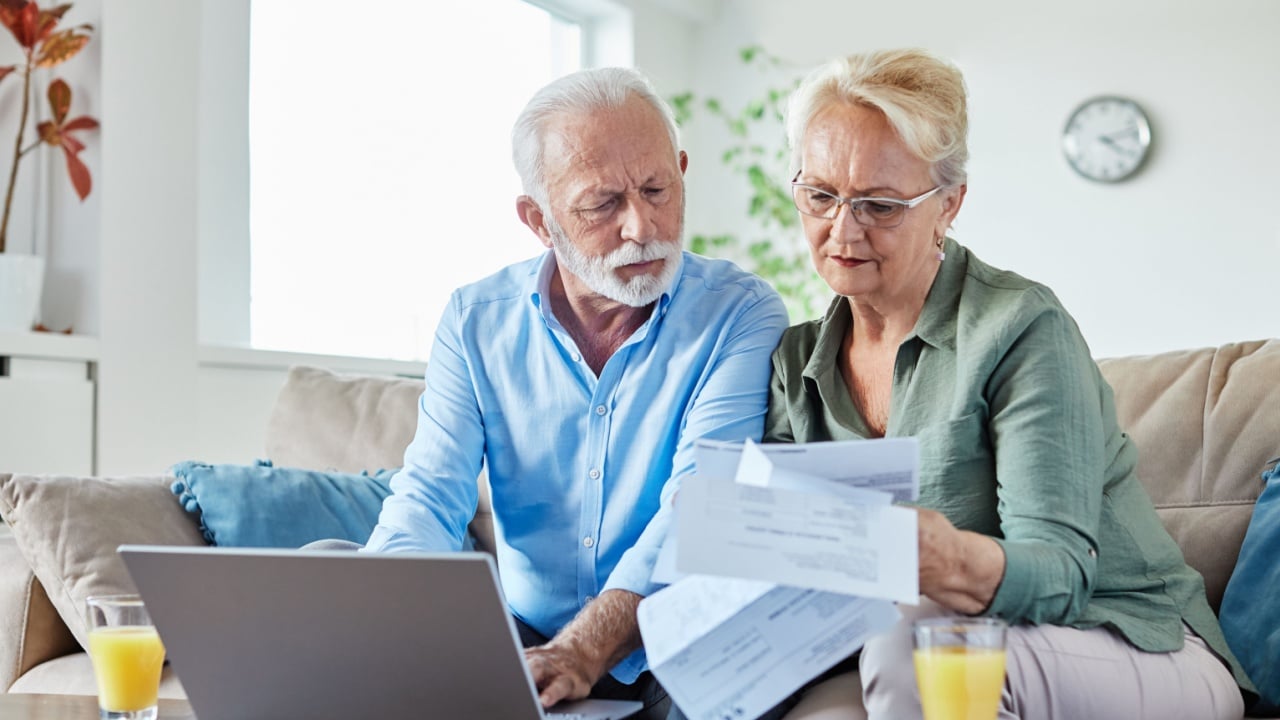 Senior couple checking bills and calculating expenses using a laptop at home, debt taxes concept, worried married elderly couple