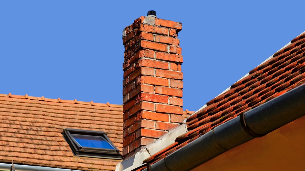 Isolated damaged clay brick chimney with weathered and spalling surface. clay tile roofing. metal flashing. strong shadow. blue sky background, summer scene.