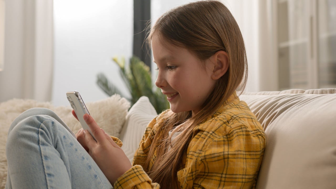 Smiling carefree little Caucasian kid girl scrolling typing screen playing games online using mobile phone telephone application. Child daughter schoolgirl browsing Internet couch home. Device overuse