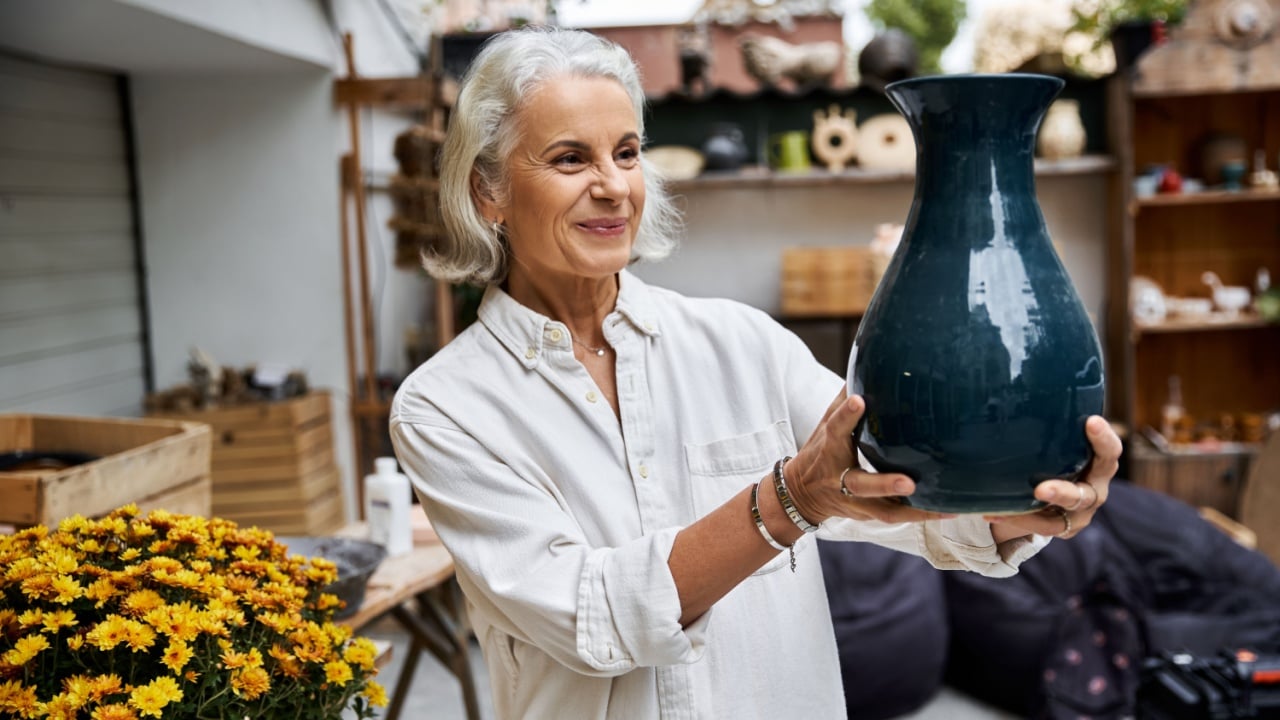 A joyful mature woman presents a striking pottery vase surrounded by lush greenery.