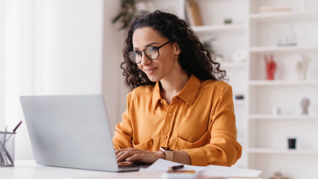 Middle Eastern Lady Using Laptop Working Online Wearing Eyewear Sitting At Workplace In Modern Office. Remote Job, Technology And Career Profession Concept. Side View