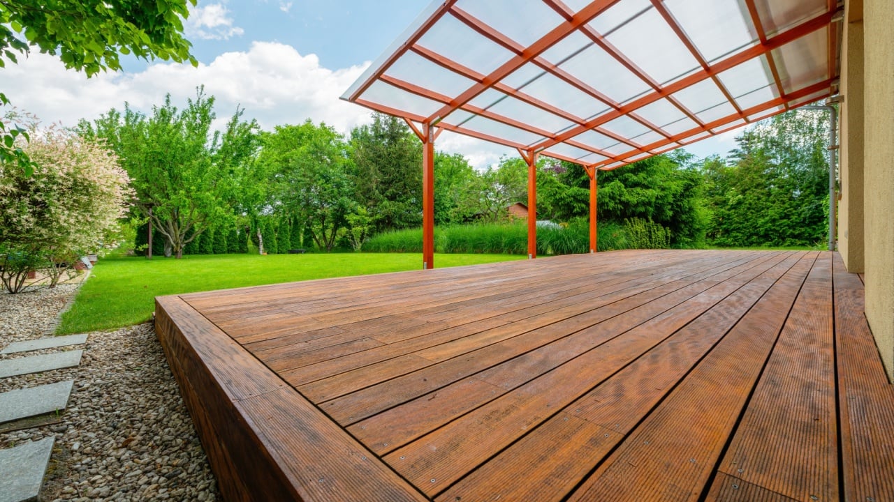 Wooden garden deck with a polycarbonate pergola roof, surrounded by a lush green backyard and landscaping, providing a shaded outdoor relaxation area