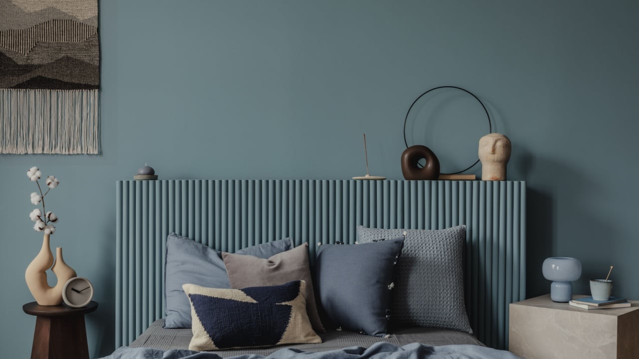 A close-up of a minimalist bedside setup featuring cotton stems in a sculptural vase, a sleek blue lamp, and stacked books for a peaceful reading corner.