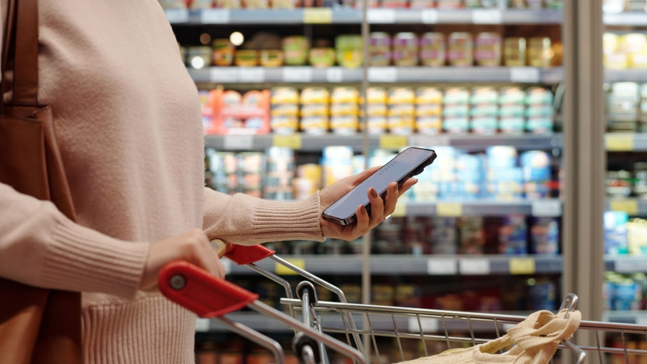 Side view of unrecognizable adult woman doing grocery shopping in supermarket and using phone while pushing cart, copy space