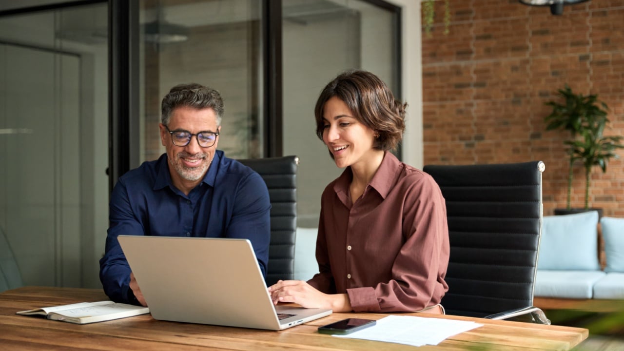 Female manager showing financial results to colleague at work. Two professional team business man and woman using laptop at corporate meeting. Happy busy executive people working together in office.