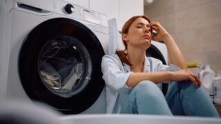 Exhausted woman waiting for washing machine to finish in laundry room.