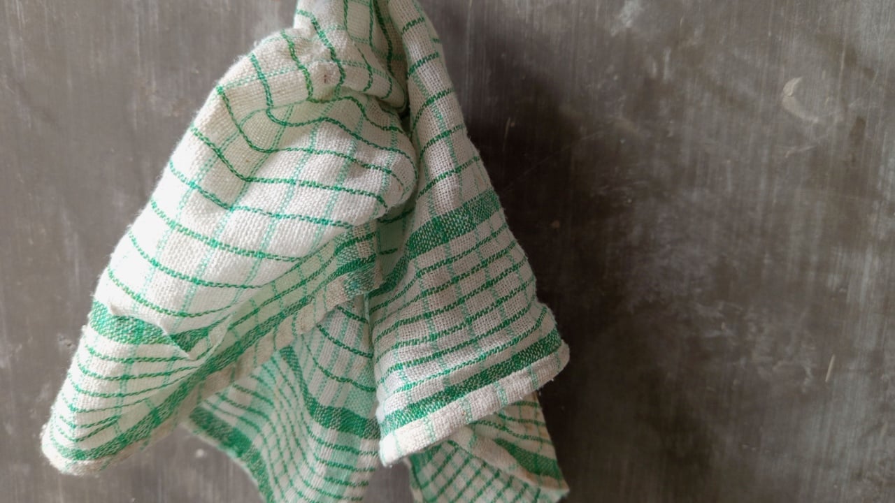 A hand holding a white kitchen towel with green checkered patterns. The towel appears slightly worn and wrinkled, suggesting regular use. The background is a plain, textured gray wall.