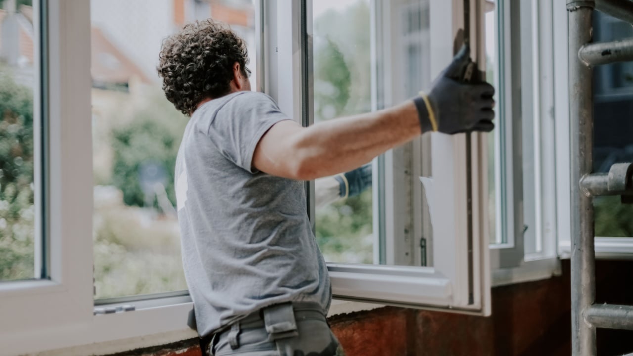 One young handsome Caucasian man in a gray uniform stands from the back and repairs installing a window frame in a house, close-up side view. Concept for home renovation and window installation.