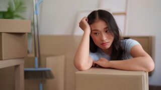 Young adult Asian female experiences moving day fatigue surrounded by relocation packing boxes ready for transport or unpacking in new apartment residence