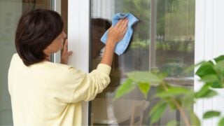 Mature woman washing glass door with blue microfiber cloth, indoors. Cleaning and professional house cleaning, small business