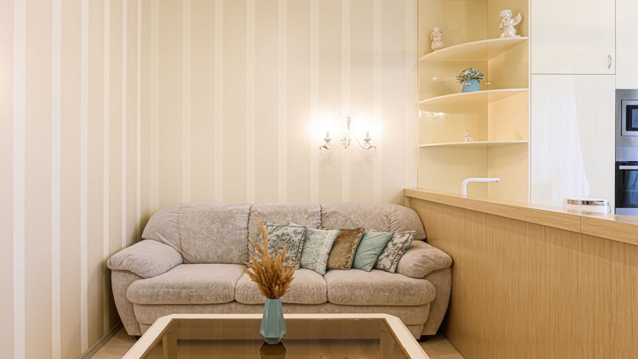 Cozy living space features a beige sofa with decorative pillows, a glass-topped table, and a stylish built-in shelf. Vertical striped wallpaper adds subtle charm
