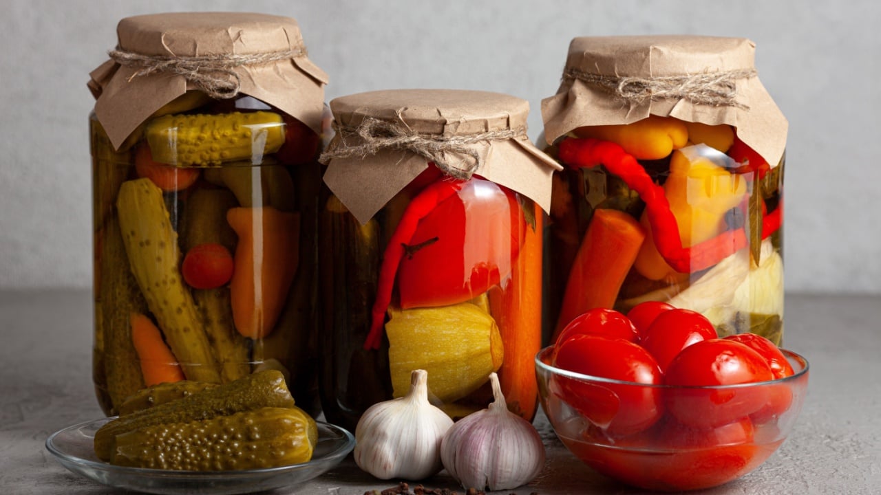 Three jars filled with a vibrant mix of pickled vegetables, sealed with kraft paper and twine. Surrounded by garlic, black pepper, and a bowl of juicy red tomatoes.