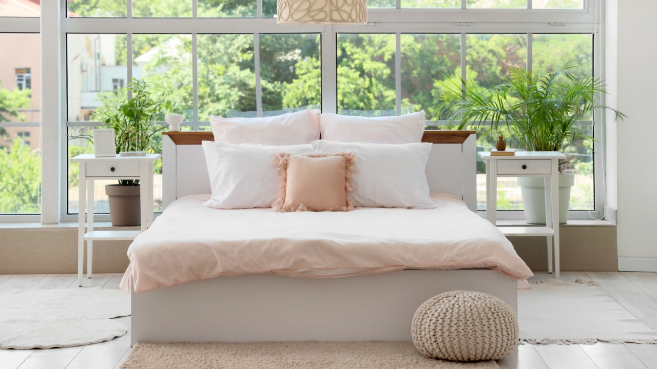 Bed with stylish pink bedding, bedside tables, ottoman and houseplants near window in bedroom