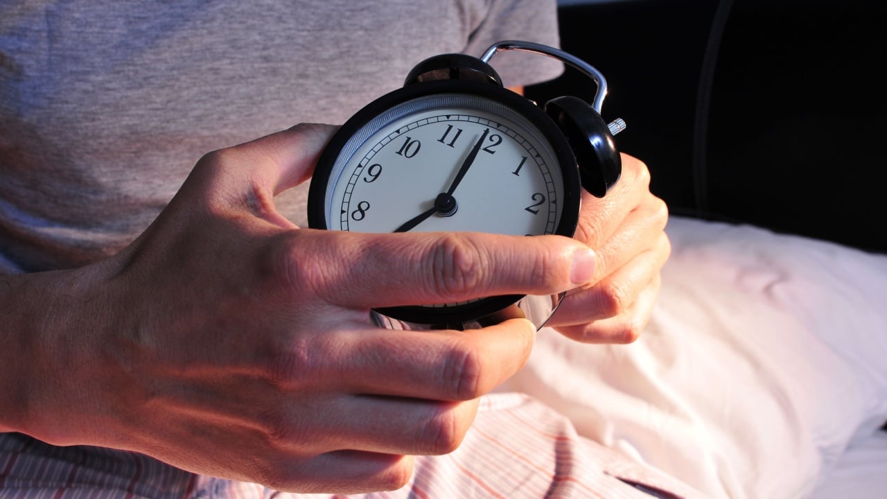 a young caucasian man wearing pajamas in bed setting the alarm clock at 7 before lie down