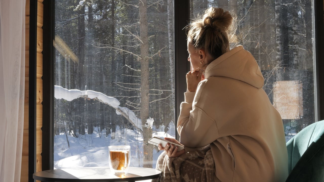 Woman wrapped in warmth enjoys quiet winter day looking out window feeling peaceful in soft light from window lost in thought by window while snow lies outside moment of quiet comfort and reflection