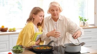 Happy senior woman preparing soup and vegetables while teaching her granddaughter cooking with recipe book in kitchen