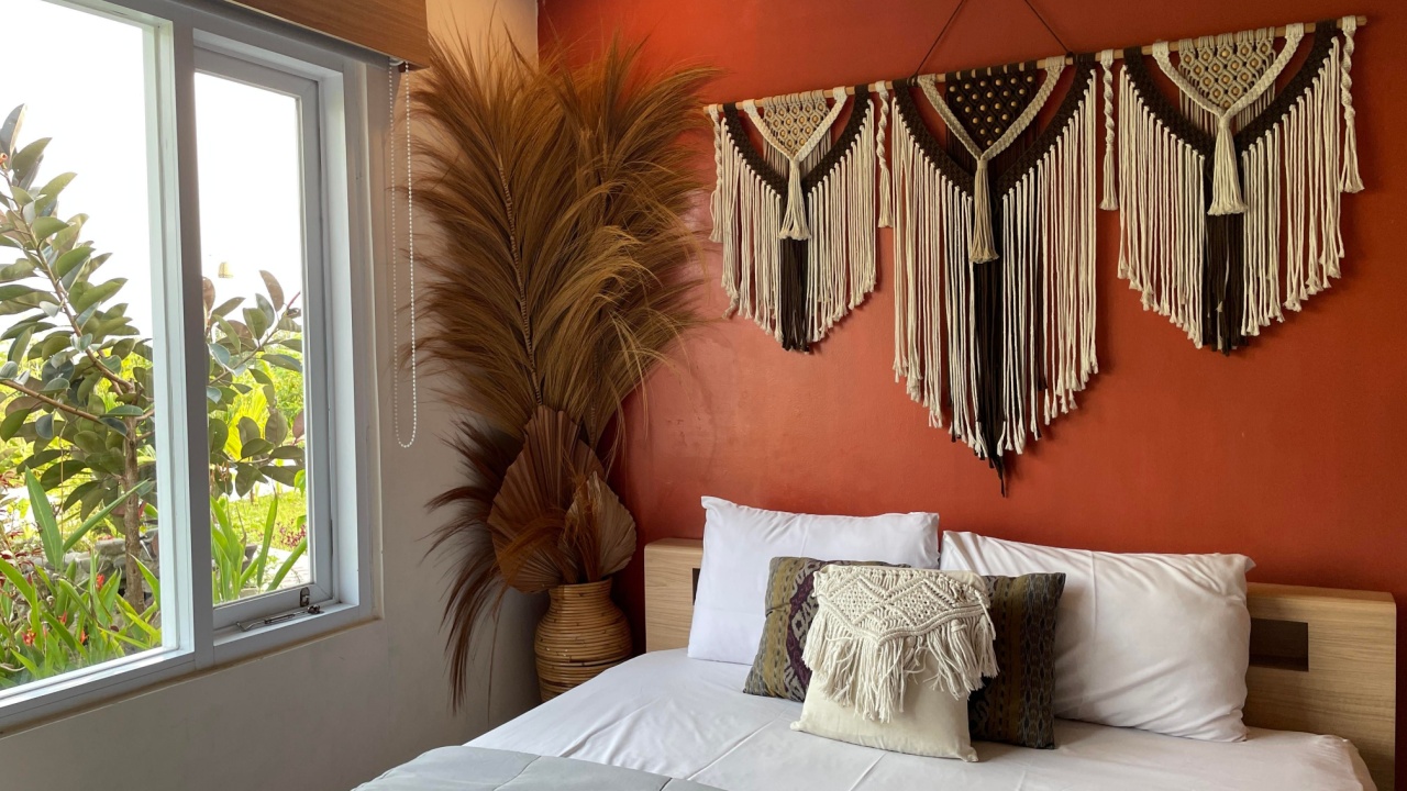 A cozy and stylish bedroom featuring a warm red accent wall decorated with intricate macram&eacute; wall hangings. The bed is neatly made with white linens and a grey throw, flanked by a large dried pampas grass arrangement in a woven vase near a bright window.