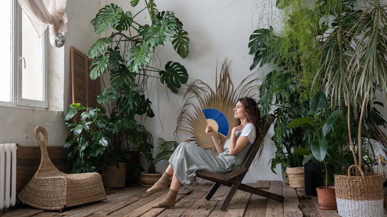 Indoor tropical oasis. Young relaxed dreamy woman using paper fan while escaping summer heat in beautiful and refreshing home garden full of large-leaved exotic plants, dreaming about vacation