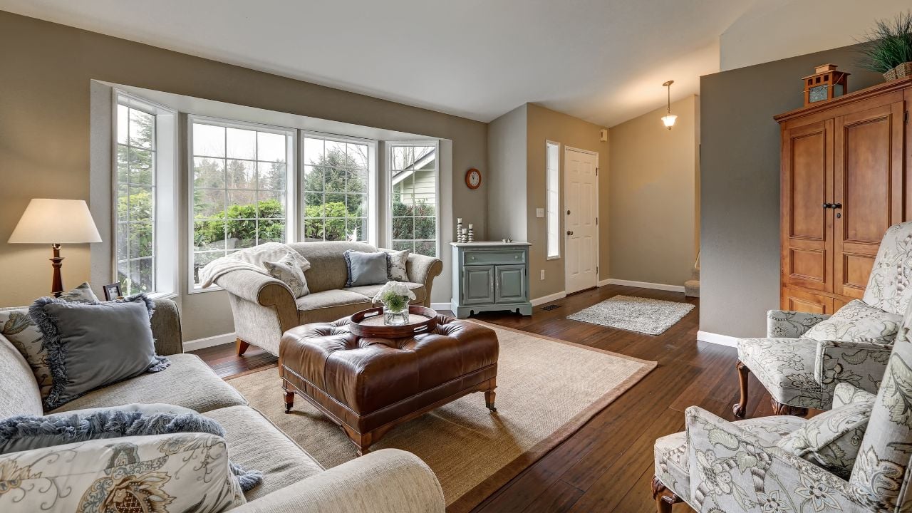 Cozy family room interior with taupe walls, sloped ceiling and wide plank wood floors. The room is furnished with claw foot chairs facing comfortable sofas across from brown leather tufted ottoman.