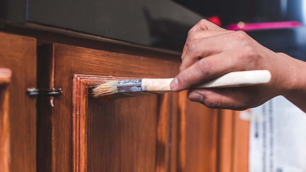 A handyman paints a fresh coat of varnish on the surface of a base kitchen cabinet with a medium sized brush. Home renovation or finishing works.