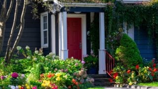 A red door on a wooden navy blue house with white trim. The cottage has lush greenery around the entrance: red, yellow, pink, and white colored flowers, hydrangea shrubs, and green grass in front.