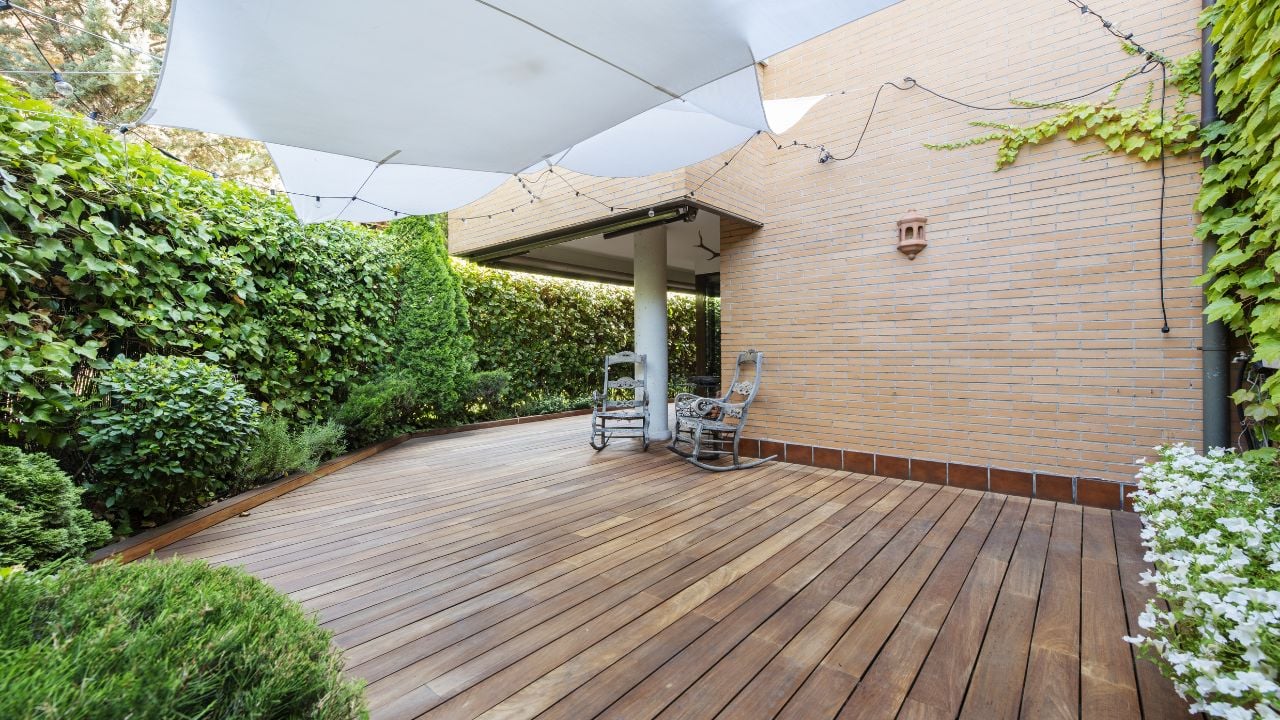 Terrace of a ground floor house with hedges and ornamental plants, slatted acacia hardwood floors twin wooden sunbeds and white awnings 
