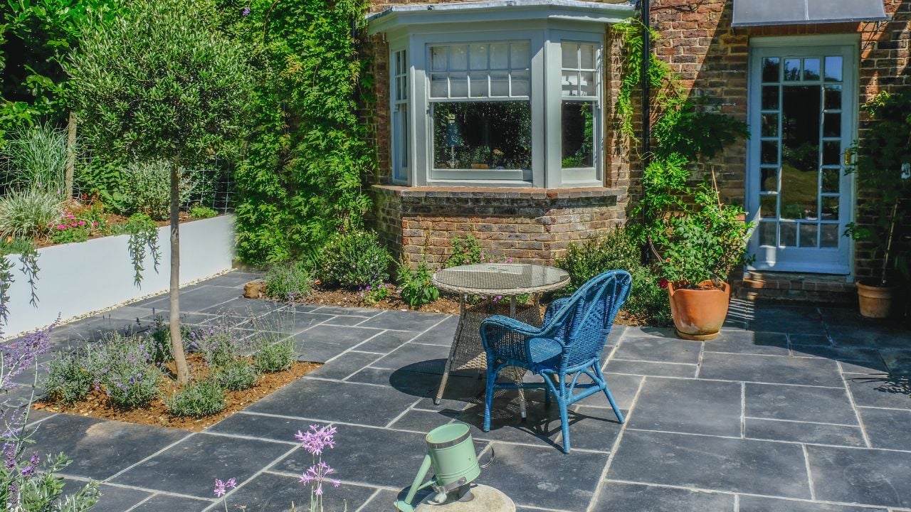Blue wicker chair and wicker table in the middle of a slate patio with plants and trees in beds and pots. Taken on a sunny summer day with nobody in the shot. 