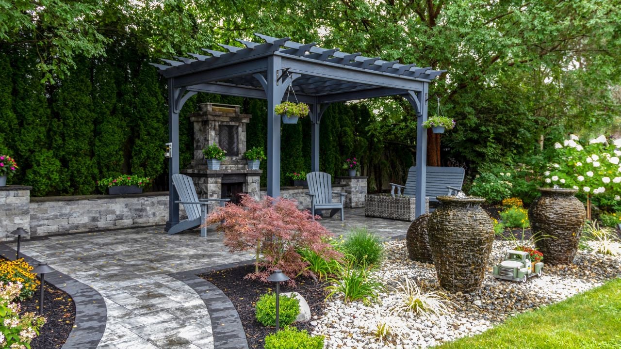 Large gray pergola on pavers with stone fireplace and waterfall urns for backyard oasis landscape design 