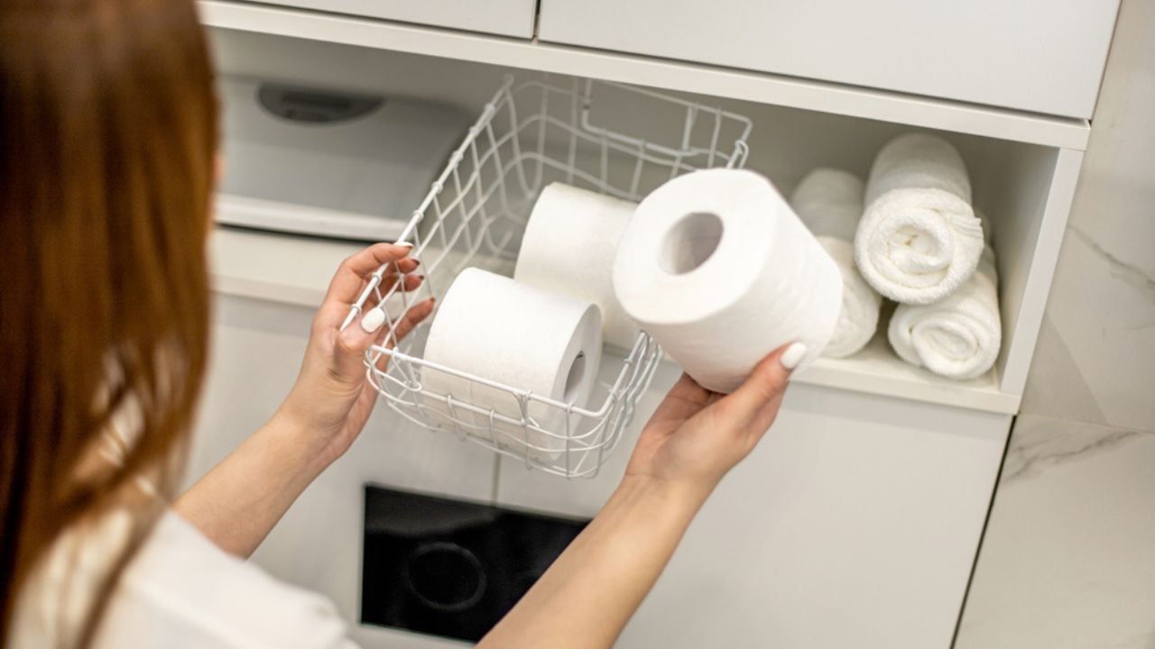 Young woman is organizing and placing mesh basket with stack of toilet paper rolls in bathroom cupboard near rolled up hand towels.